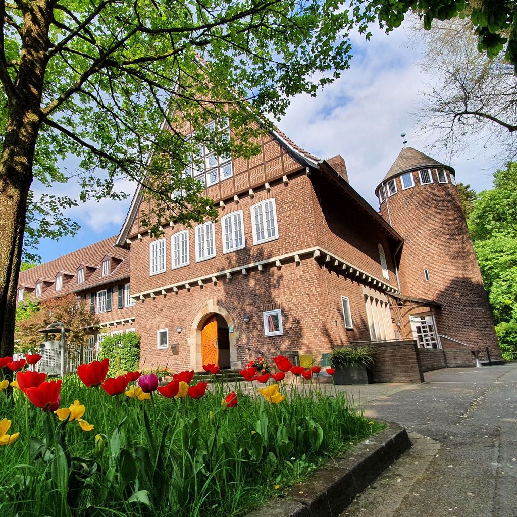 Alt-Text: Historisches Backsteingebäude mit Turm und blühendem Tulpenbeet vor grünem Baum.