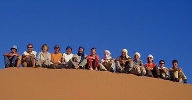13 Männer und Frau sitzen auf einer Wüsten-Düne vor einem strahlend blauen Himmel. Sie sind wüstentauglich gekleidet.