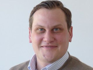Lächelnder Mann mit Brille, grauer Jacke und gestreiftem Hemd.