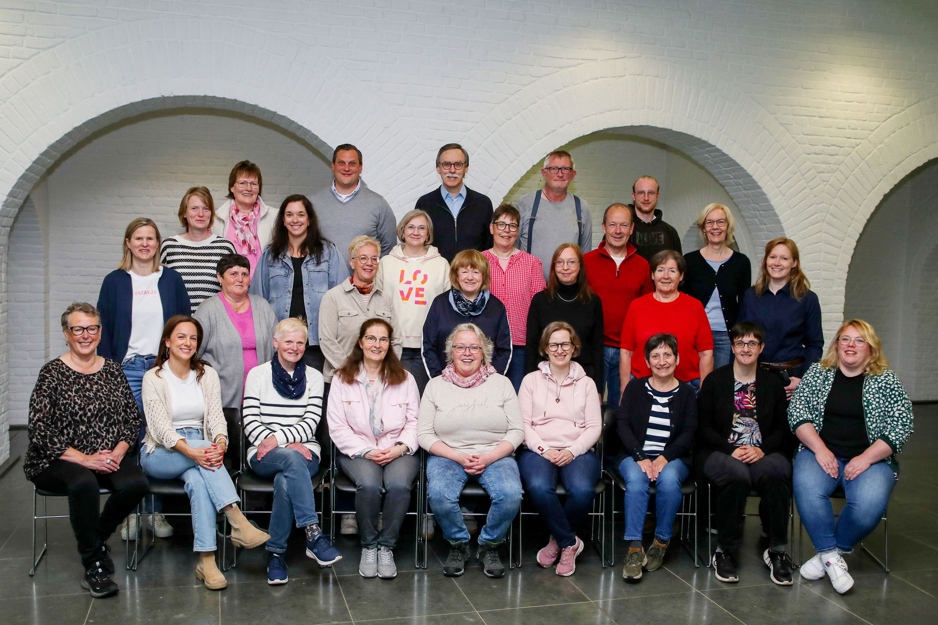 Großgruppe von 20 Personen, lächelnd im Gruppenfoto vor weißer Wand mit Nischen.