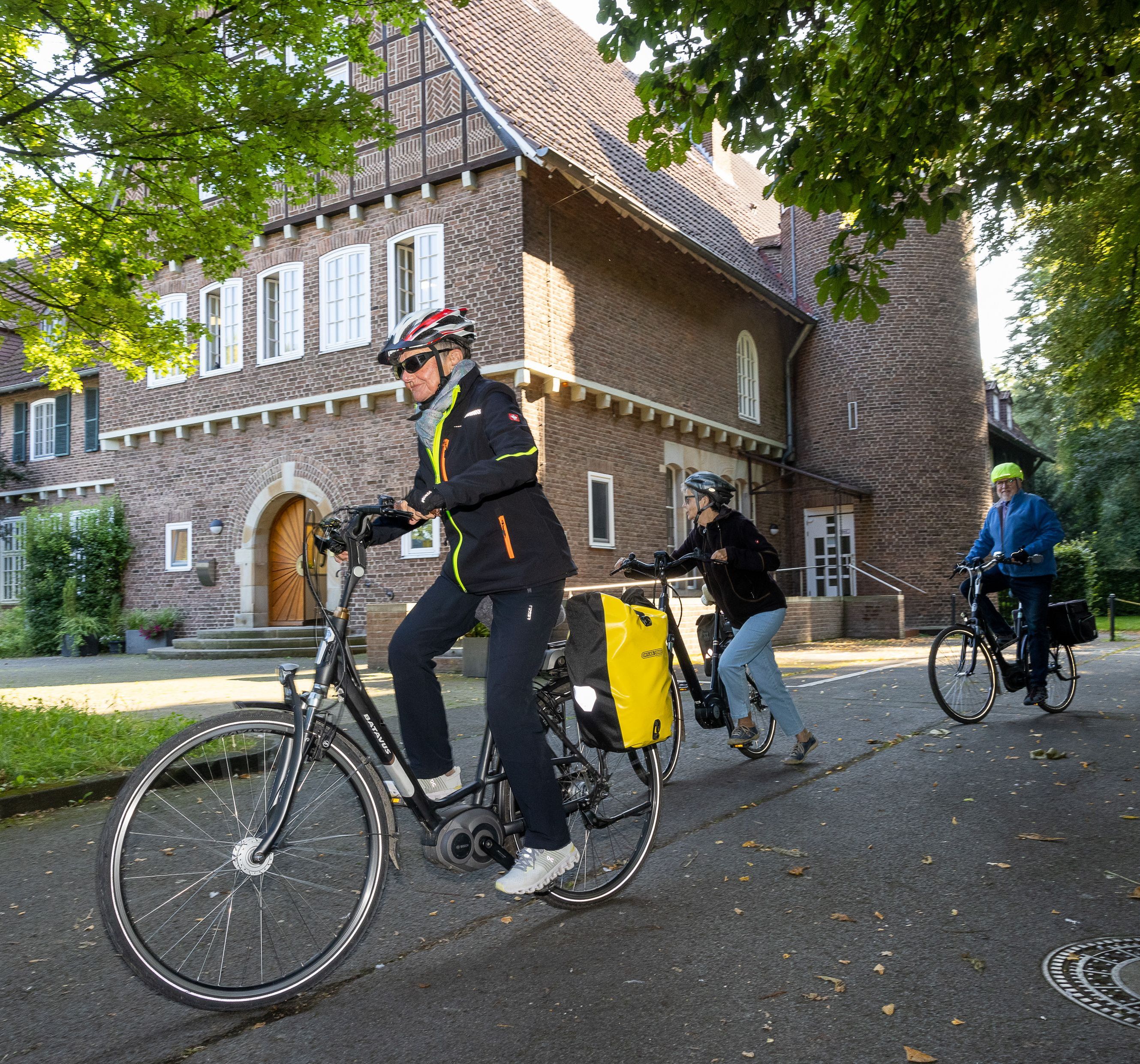 Drei Radfahrer mit Helm und Gepäck fahren auf einer Straße vor historischem Gebäude vorbei.