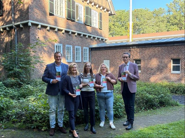 Fünf Personen halten Diplome vor historischem Backsteingebäude im Garten.