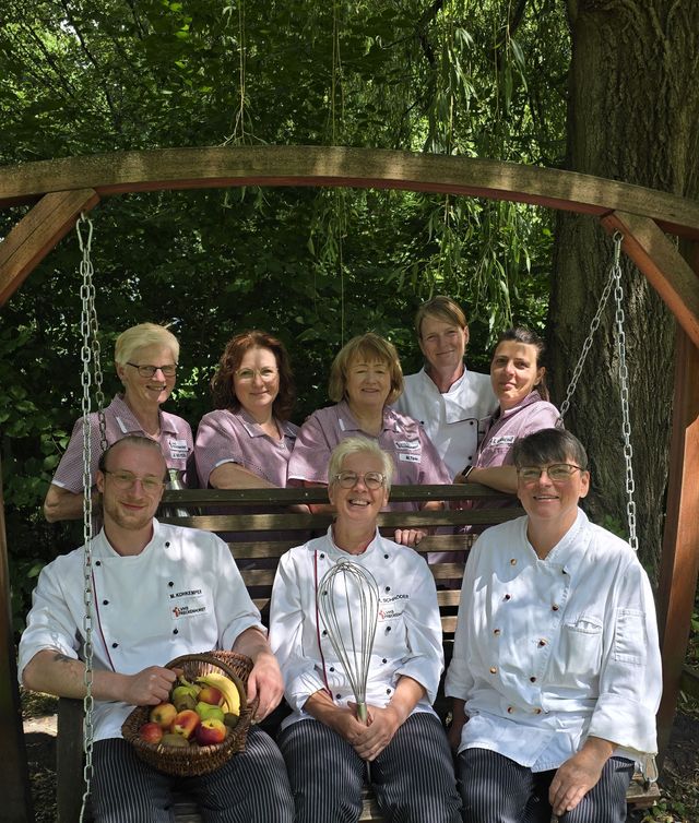 Glückliche Köche und Gäste auf Holzbank unter Baumschaukel mit Obstkorb.