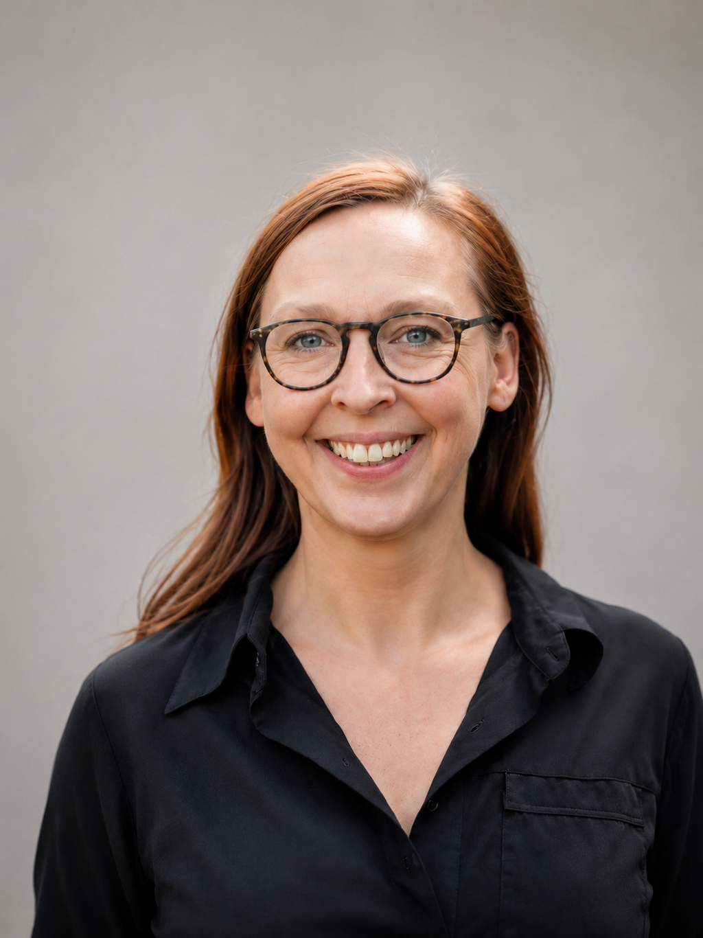 Lächelnde Person mit roten Haaren, Brille und schwarzem Oberteil vor hellem Hintergrund.