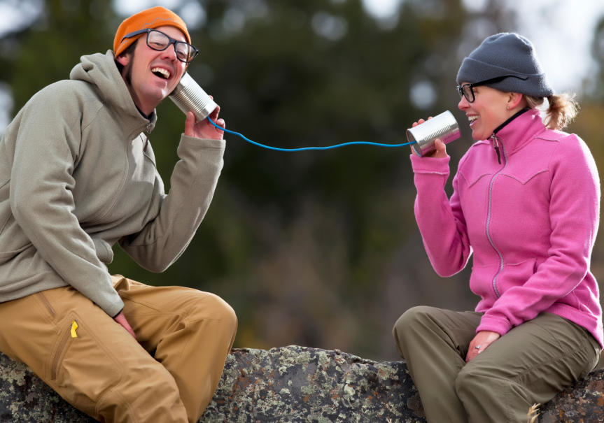 Ein Mann und eine Frau winterlich gekleidet sitzen auf einem großen Stein. Sie haben jeder eine Blechdose in der Hand die mit einer Schnur verbunden sind. Die Frau spricht in die Blechdose der Mann hält sich die Blechdose ans Ohr 