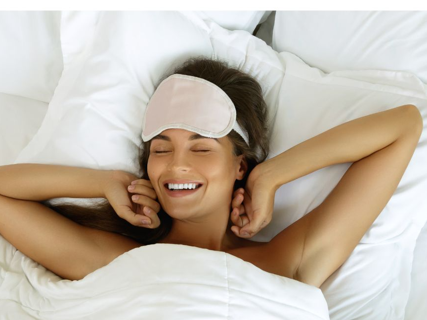 Junge Frau mit braunen langen Haaren liegt in einem weißen Bett und räkelt sich lächelnd. Auf ihrem Kopf trägt sie eine Schlafmaske
